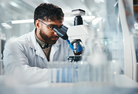 Medical environment with a scientist in a laboratory looking through a microscope with engineering plastics and test tubes in the foreground